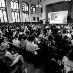A black and white image of a packed audience at IMC 2019 keynote lecture.