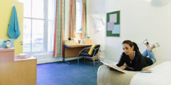 A room in Lyddon Hall showing a female student reading on a bed with a desk and window behind