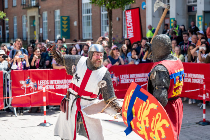 Two re-enactors in medieval armour carrying swords and shields demonstrate medieval combat techniques in front of a packed crowd in an outdoor arena. 