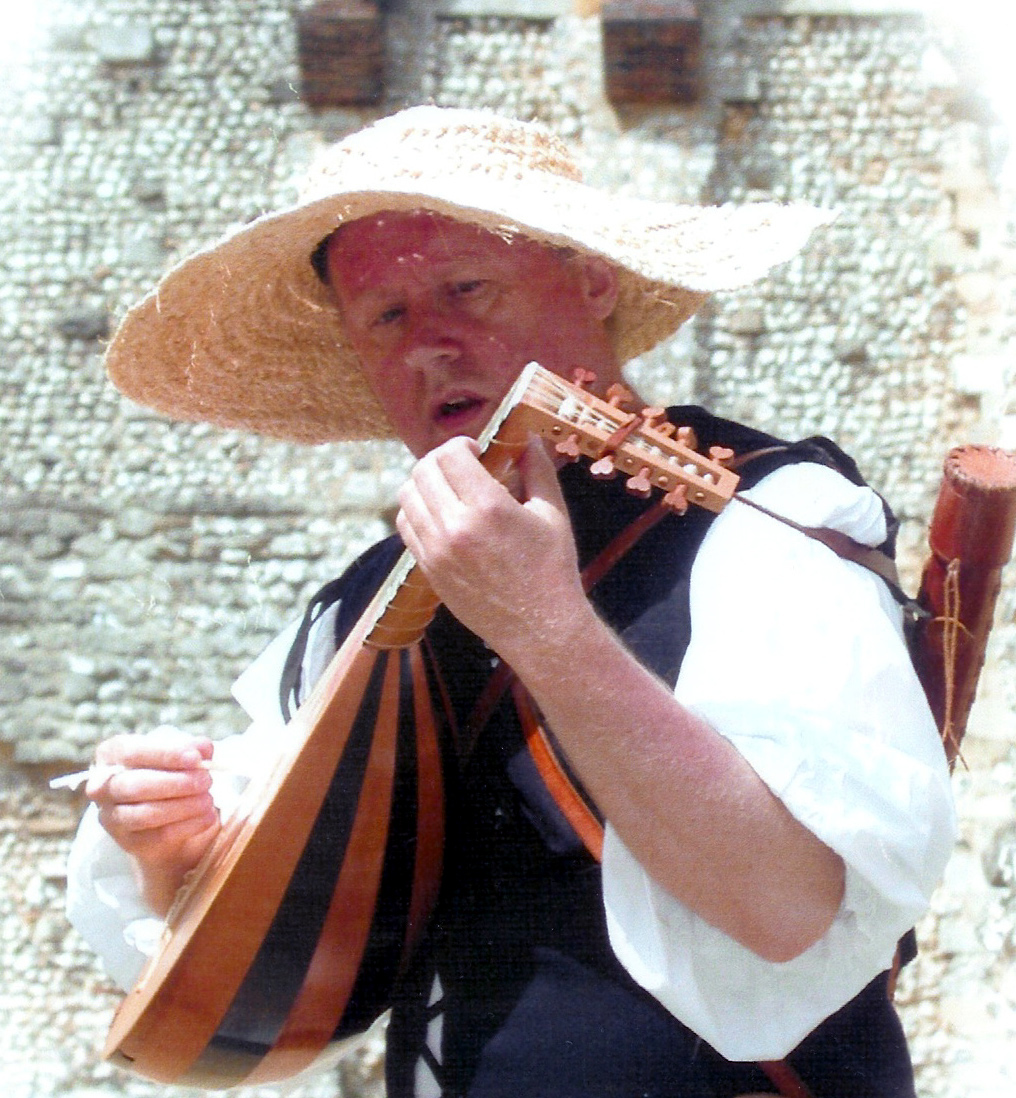 Peter Bull is wearing a straw hat, traditional medieval clothes and strumming a lute or similar medieval instrument.