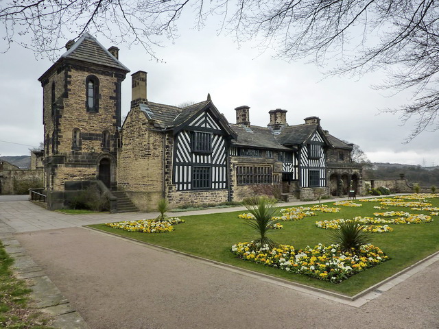 A photograph of Shibden Hall, an impressive Tudor panelled building with a tower to the left, two wide frontages and an ornamental garden in the foreground.