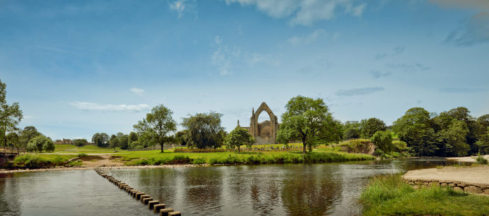 A panoramic view of Bolton Abbey ruins with a wide, brown river in the foreground.