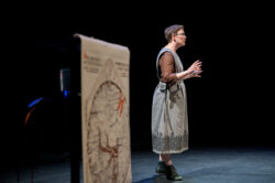 Daisy Black, a performer in a blue dress, gestures emphatically as she tells a story. In the foreground, a medieval map of the world is on display.