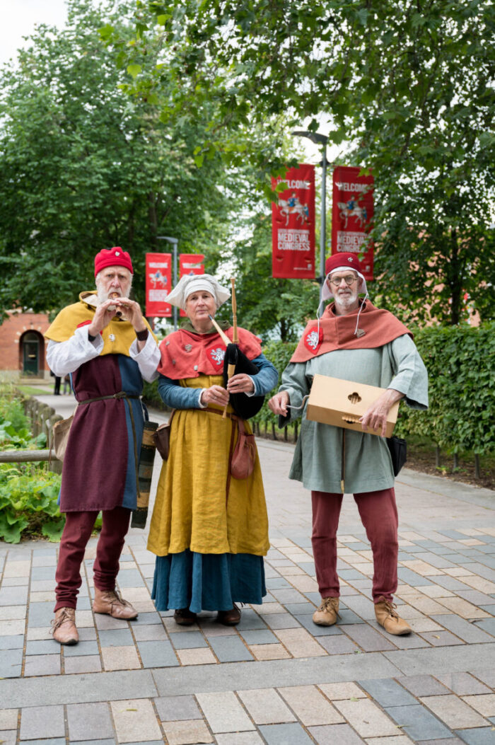 the three musicians in medieval dress stood facing the camera holding their instruments on the University of Leeds campus.