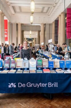 A smiling Bookfair Exhibitor stood behind their blue stall with De Gruyter Brill written on the front and books of different colours on both the table itself and on stands.